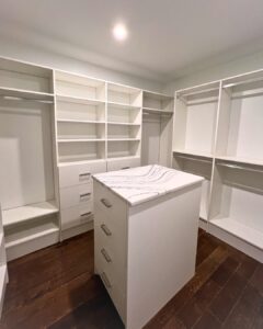 Photo of a walk-in closet in a white finish with a mix of hanging, shelving, and drawer space. There is an island with extra drawers and a marble-like finished countertop.